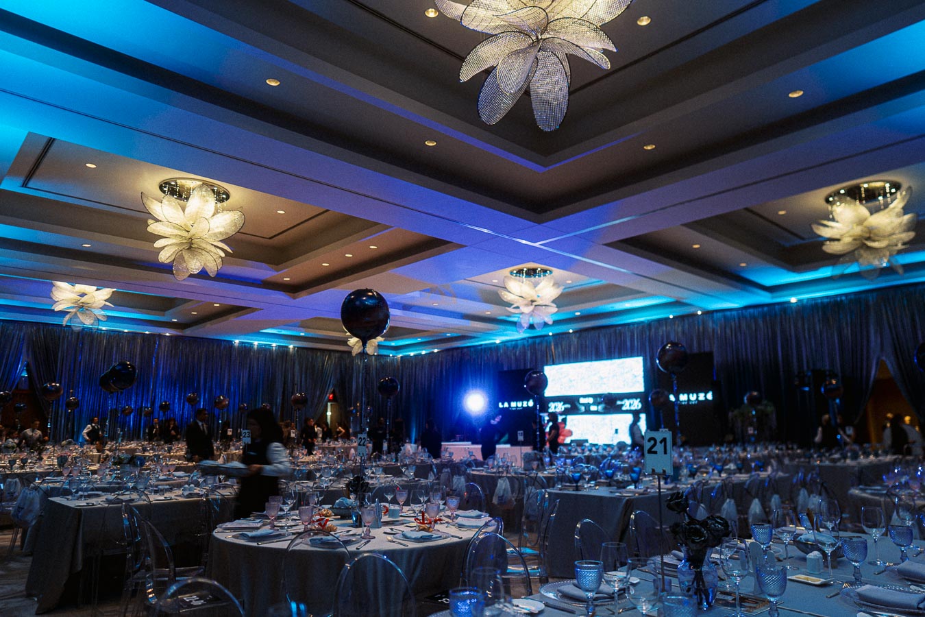 Luxurious banquet hall decorated for an evening event, featuring elegant blue lighting, opulent chandeliers, round tables set with fine glassware, and a large digital screen in the background.