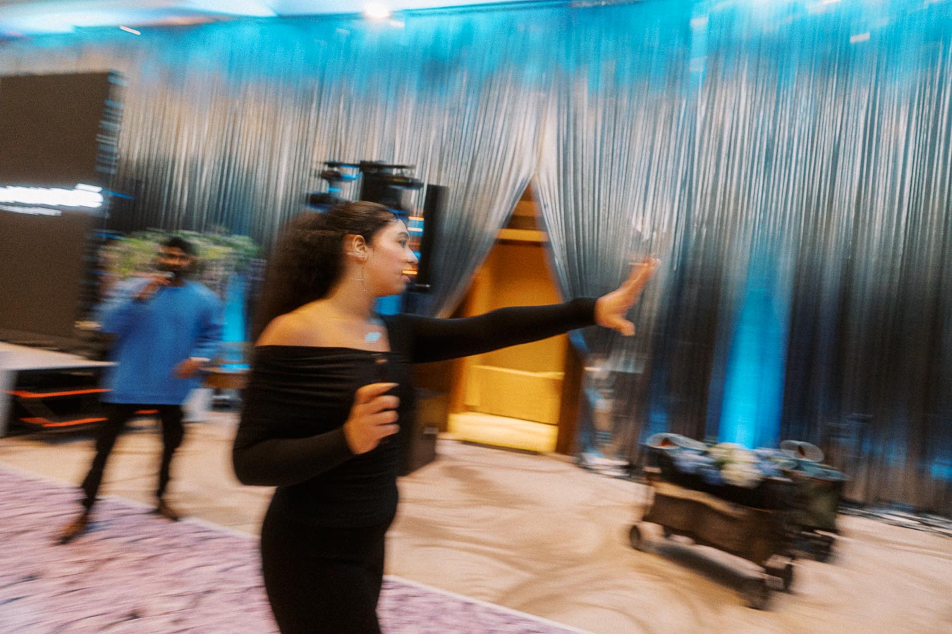 A woman in a black off-shoulder outfit gestures with her hand on a stage set with blue curtains and elegant lighting, suggesting a lively event atmosphere. A man in a blue shirt holds a microphone in the background, enhancing the dynamic scene.