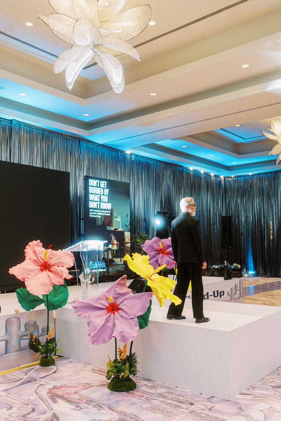 Man in suit walking on a decorated stage with large artificial flowers, under a modern ceiling light fixture, and a backdrop with a motivational message.