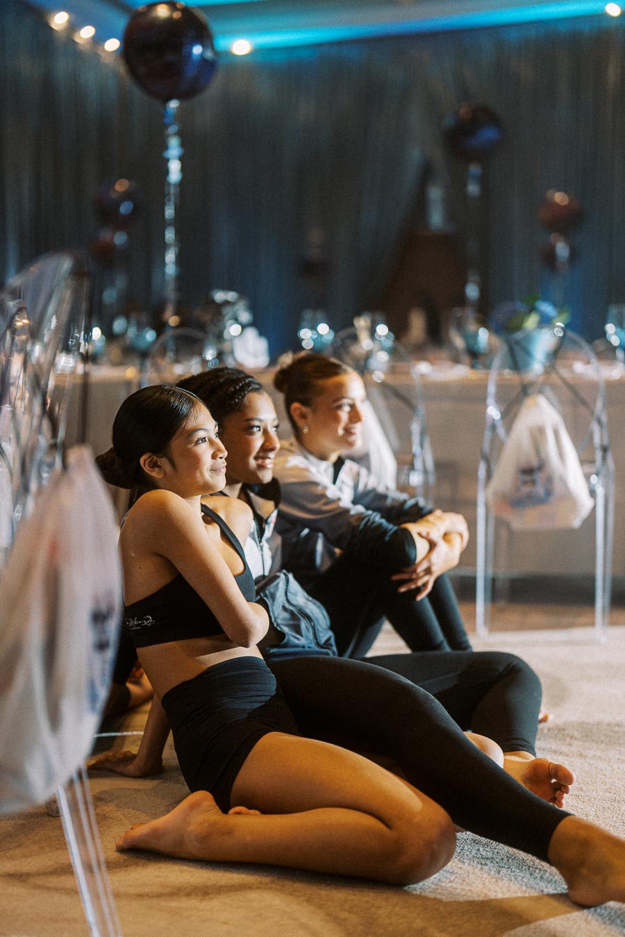 Young women sitting on the floor at an indoor event with ambient lighting and decorative balloons in the background.