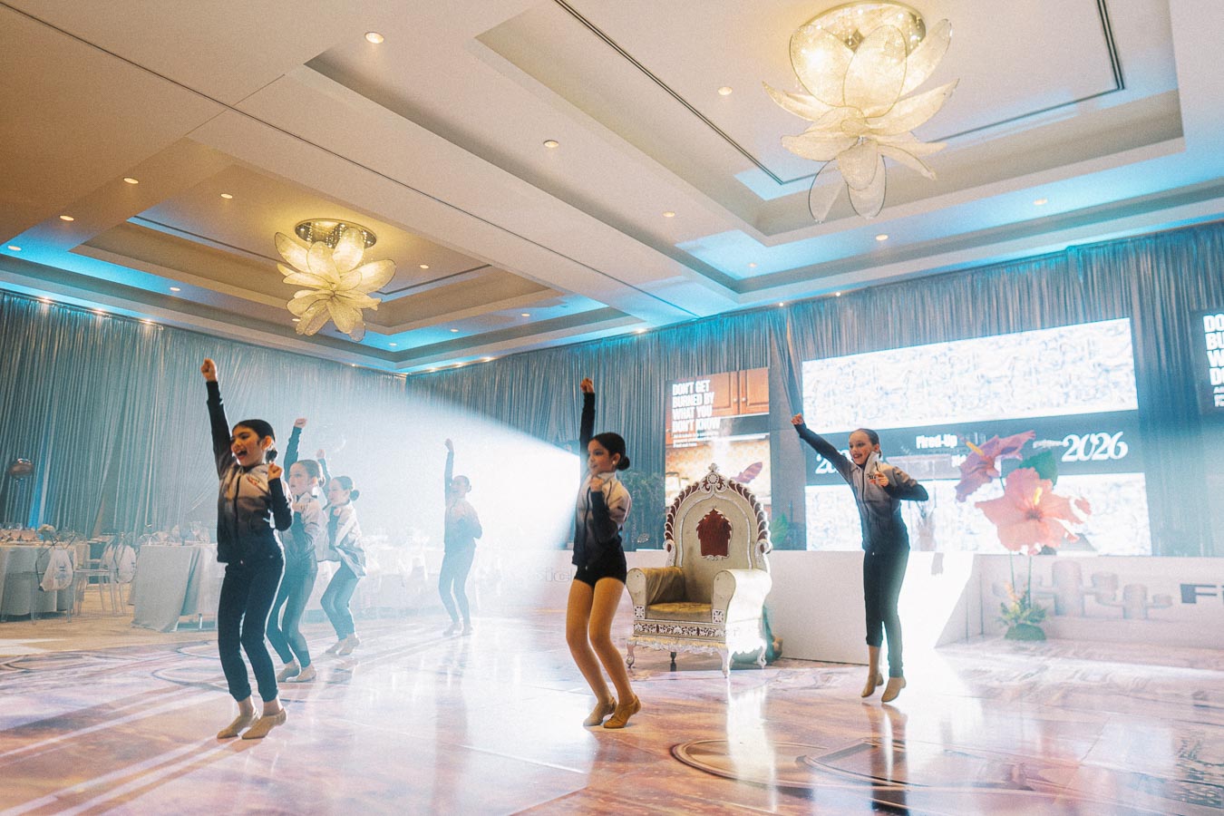 Children performing a dance routine in a beautifully decorated banquet hall with elegant chandeliers and illuminated stage backdrop.