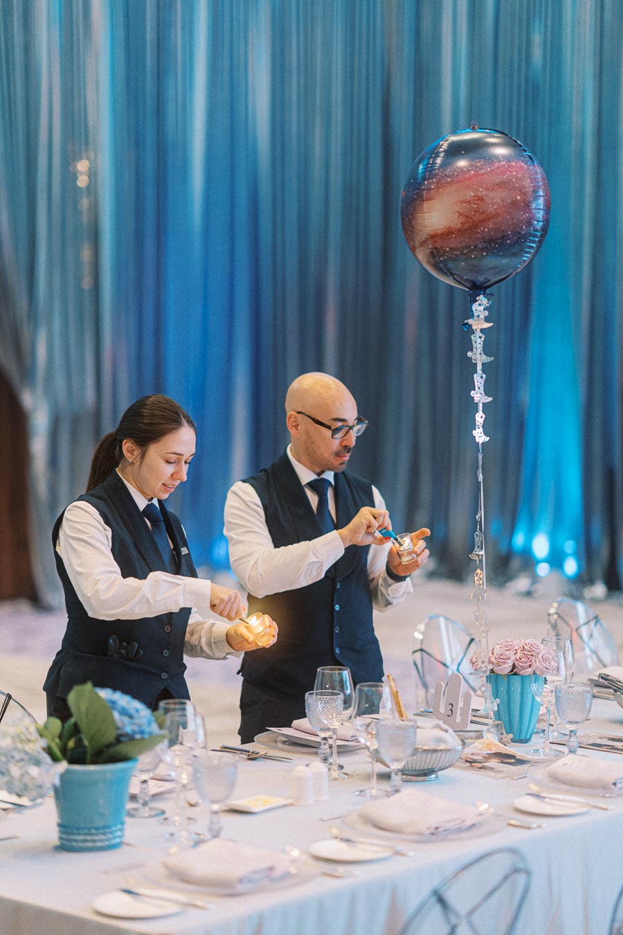 Two event staff lighting candles on a beautifully set table at an elegant event with a space-themed balloon centerpiece, against a backdrop of shimmering blue drapes.