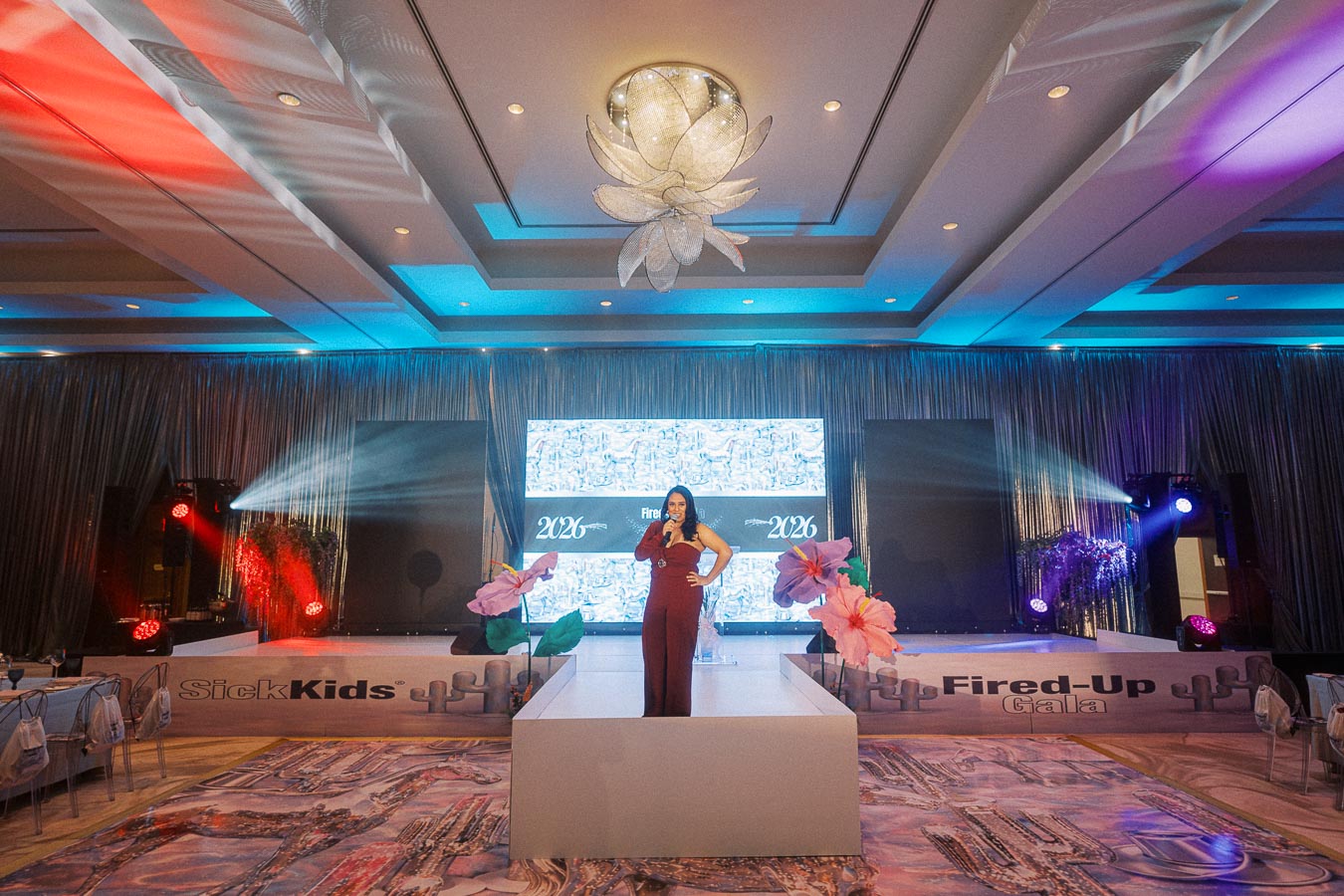 Woman speaking on stage at a luxury event gala with colorful lighting, elaborate decorations, and large flowers, featuring an LED screen background with 2026 displayed.