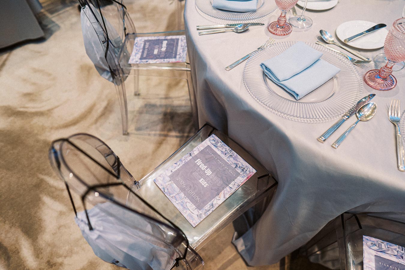 Elegant table setting with transparent chairs and a gala program, featuring pastel blue tablecloth and matching napkins. Crystal plates, silver cutlery, and pink glassware add a touch of sophistication. Suitable for upscale events or special occasions.