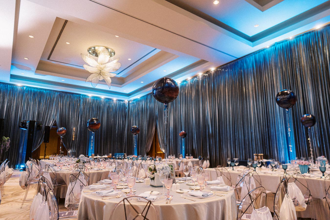 Elegant banquet hall with round tables set for a formal event, featuring blue lighting, large black balloons with branding, and modern decor.