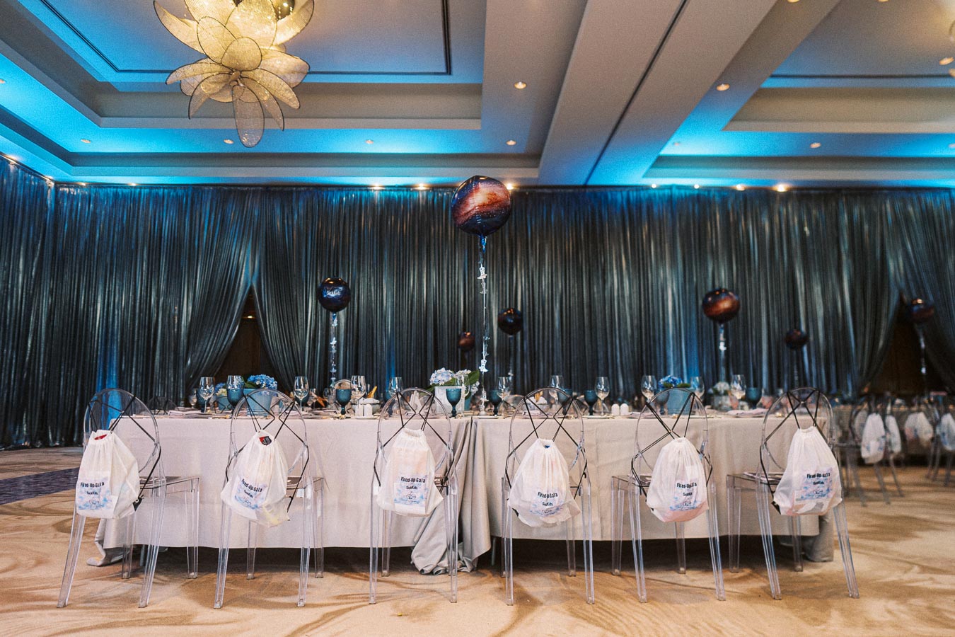 Elegant event setup with a long dining table adorned with blue accents and multiple place settings, featuring transparent chairs and decorative dark balloons under a modern chandelier.