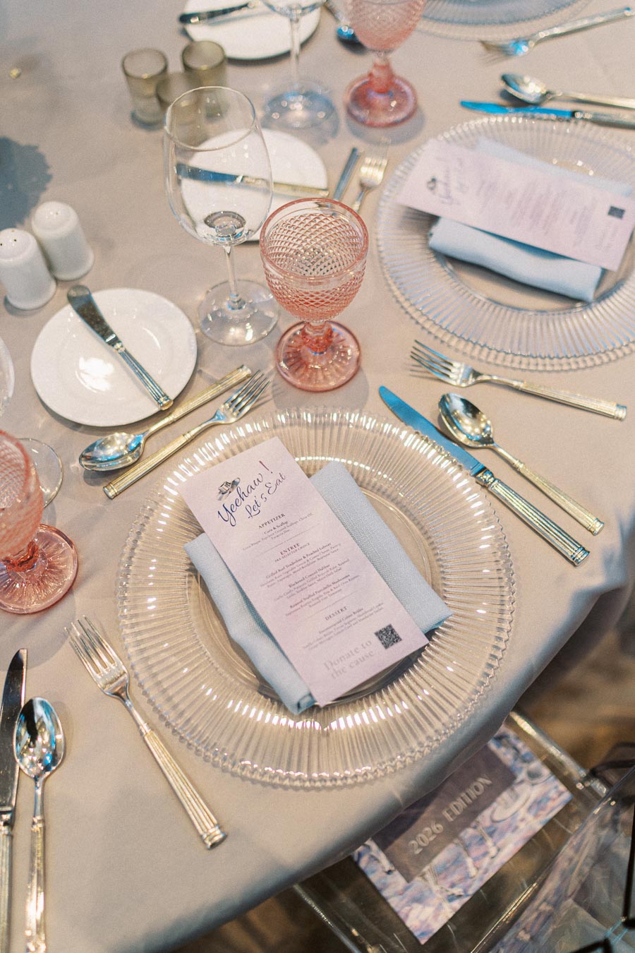Elegant table setting with fine china and glassware, featuring a detailed menu on a glass plate over a light blue napkin. The setting includes pink glass goblets, silver cutlery, and a beige tablecloth, creating a sophisticated dining atmosphere.