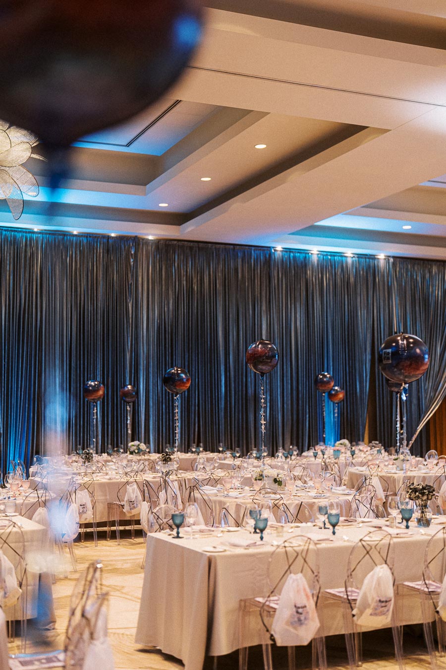Elegant banquet hall decorated with blue lighting and modern table settings, featuring transparent chairs and balloons as centerpieces, ideal for a sophisticated event or wedding reception.