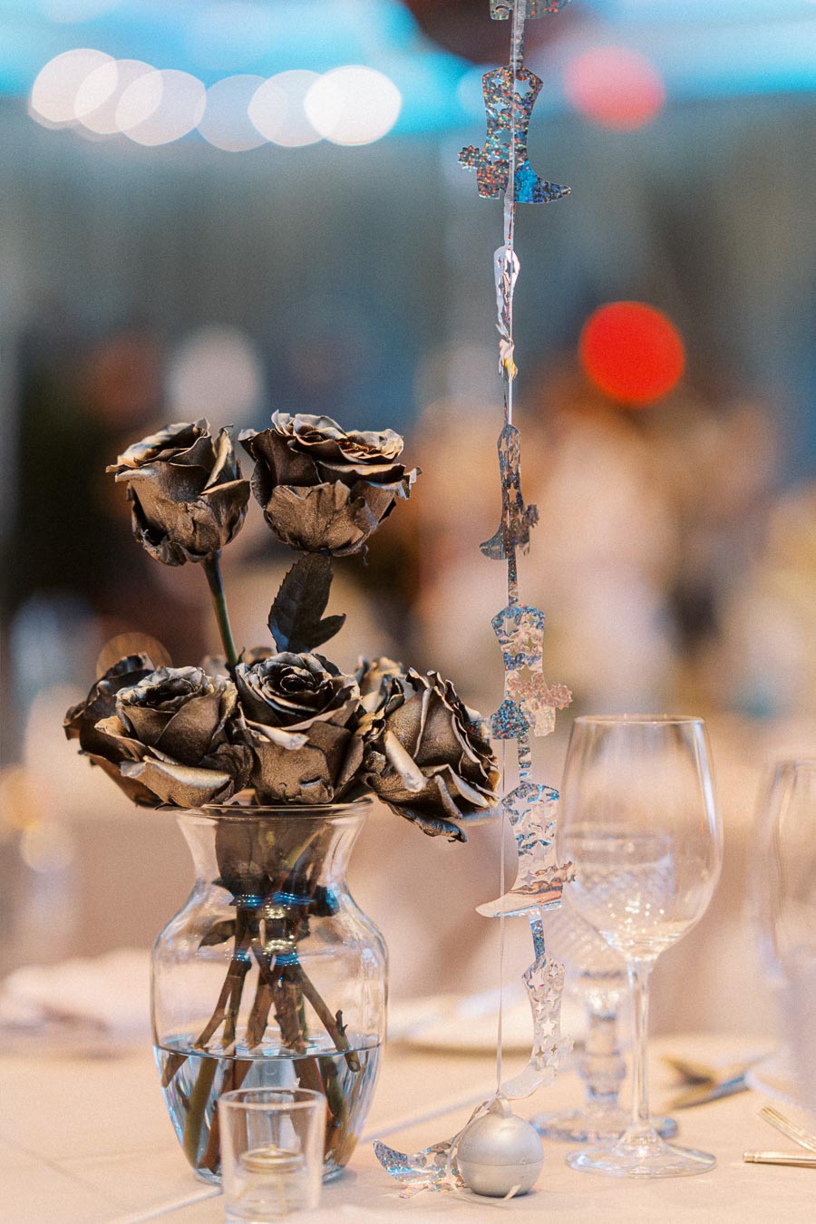 Elegant table setting featuring a glass vase of metallic-painted roses, accompanied by silver star-shaped hanging decorations, set amidst fine dining glassware for a sophisticated event ambiance.