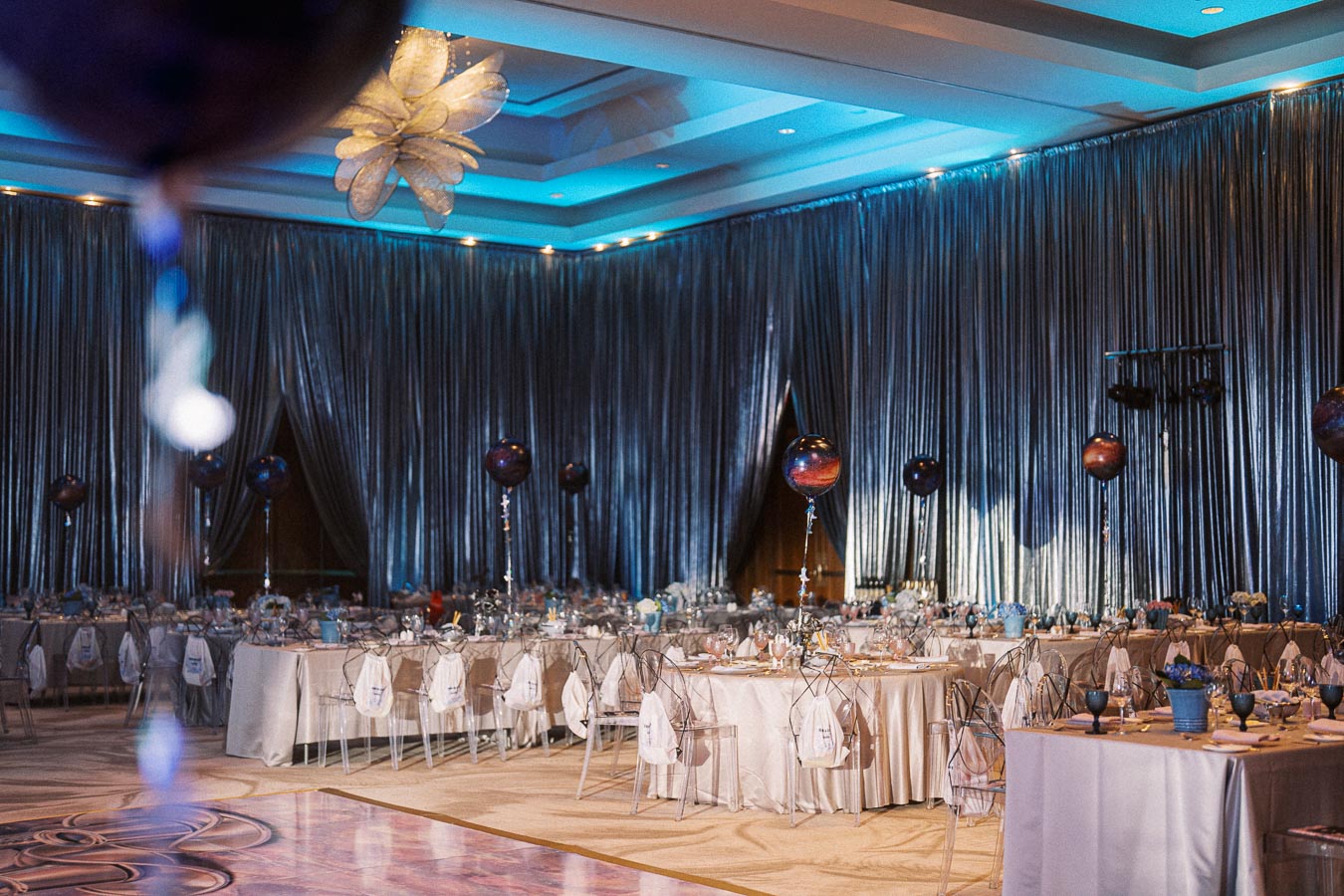Luxurious banquet hall decorated for an elegant event, featuring round tables with white tablecloths and transparent chairs, adorned with dark balloons and ambient blue lighting, creating a sophisticated and inviting atmosphere.