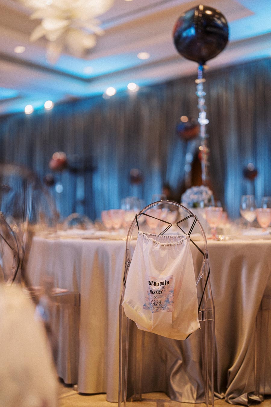 Elegant gala event setup featuring a tastefully decorated round table with a white tablecloth, glassware, and a chair adorned with a gift bag labeled “SickKids.” The background displays atmospheric lighting with blue tones and a decorative balloon centerpiece. Ideal for charity event or formal dinner presentations.