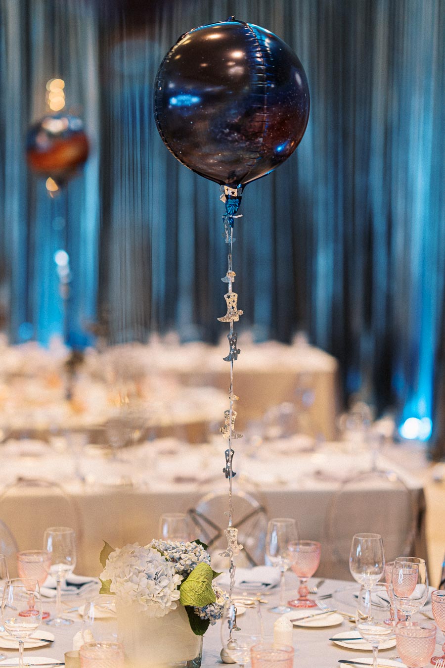 A dark starry-themed balloon centerpiece with silver star decorations and floral arrangement on an elegant dinner table setting, featuring glassware and intricate tableware, with a blurred background of a sophisticated event venue.