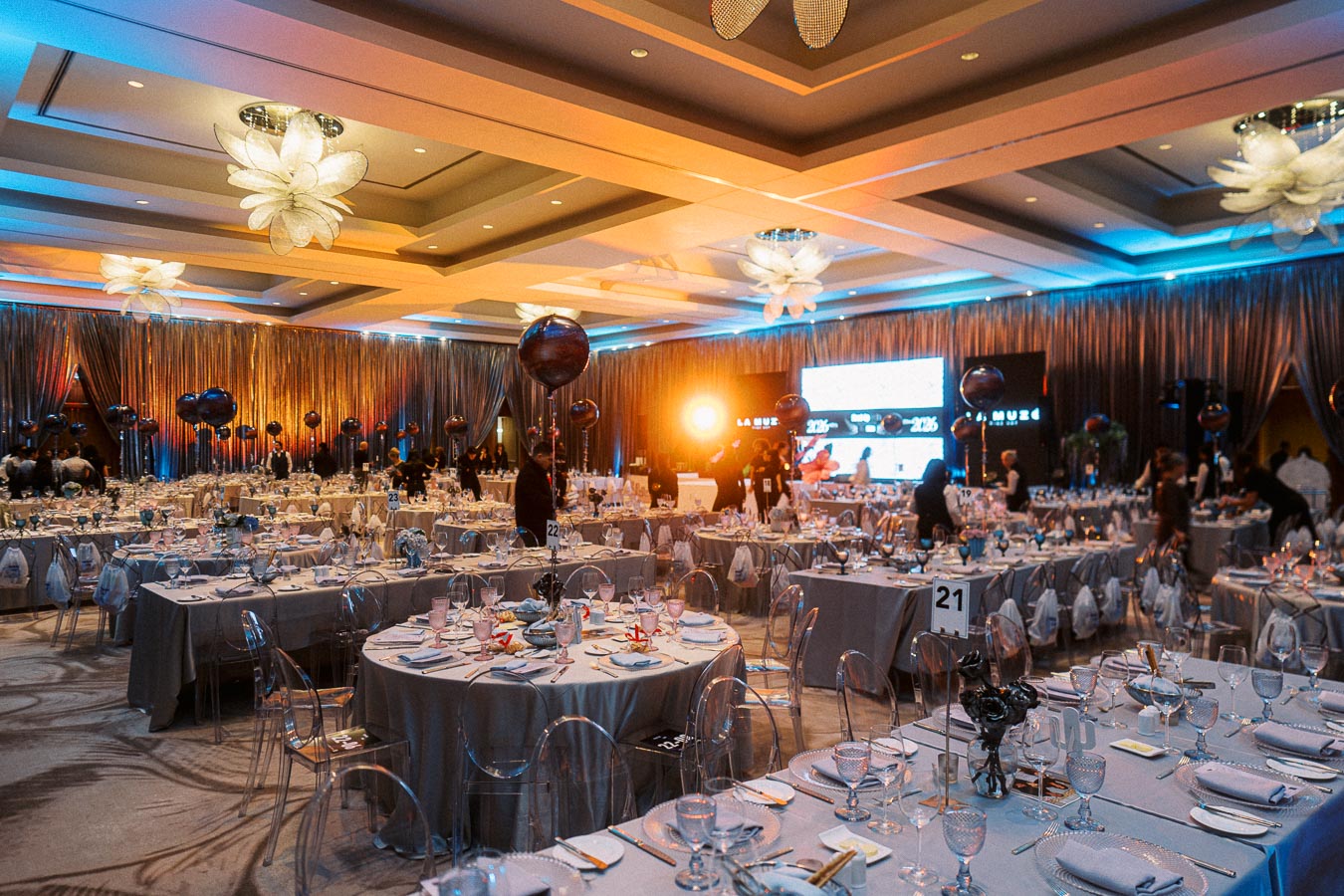 Luxurious event venue with elegantly set tables for a formal dinner, featuring stylish centerpieces, decorative lighting, and a large screen backdrop.