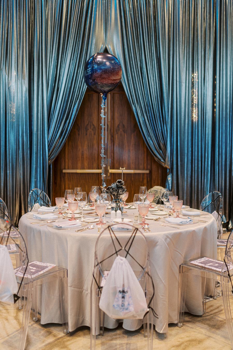 Elegant event table setup with a round table draped in a white tablecloth, adorned with glassware and folded napkins. A large, shiny balloon floats near blue, velvety curtains, creating a luxurious atmosphere. Transparent chairs encircle the table, enhancing the sophisticated decor.