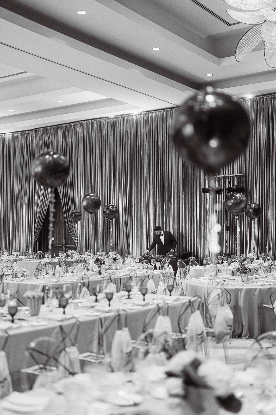 Black and white image of an elegant event space with tables set for a formal gathering, featuring chic table arrangements and decorative balloons, with a person adjusting audio equipment in the background.