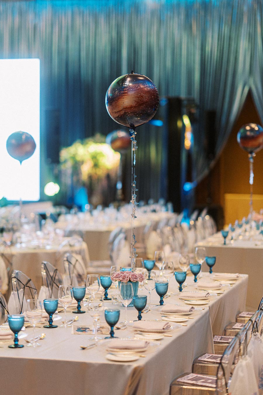 Elegant wedding reception setup featuring long tables adorned with blue goblets, rose centerpieces, and galaxy-themed balloons in a softly lit venue.