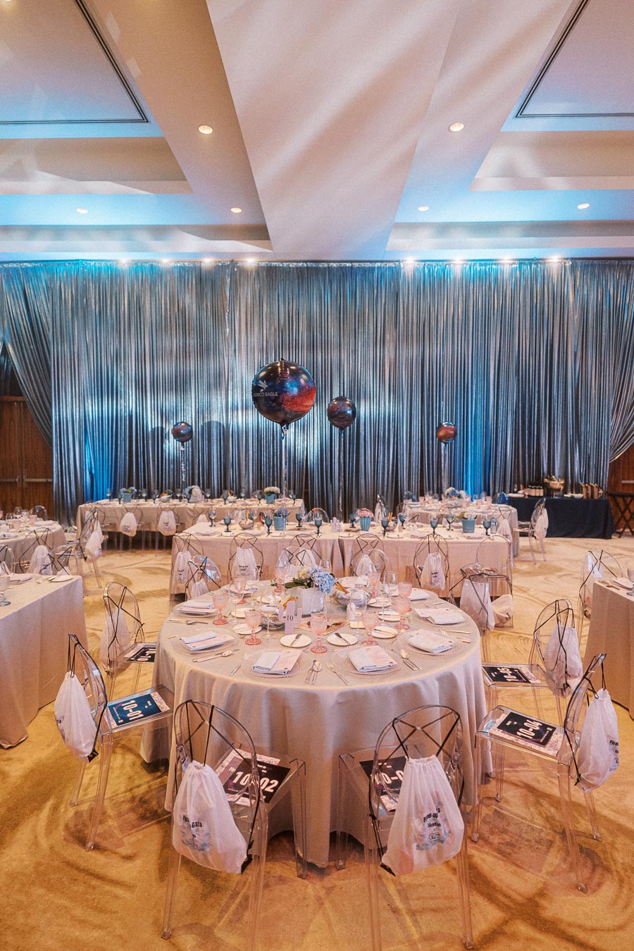 Elegant banquet hall setup with round tables covered in beige tablecloths, clear chairs, and sophisticated place settings. The space is accentuated by a shimmering silver curtain backdrop and decorative balloons, creating a refined atmosphere for a formal event.