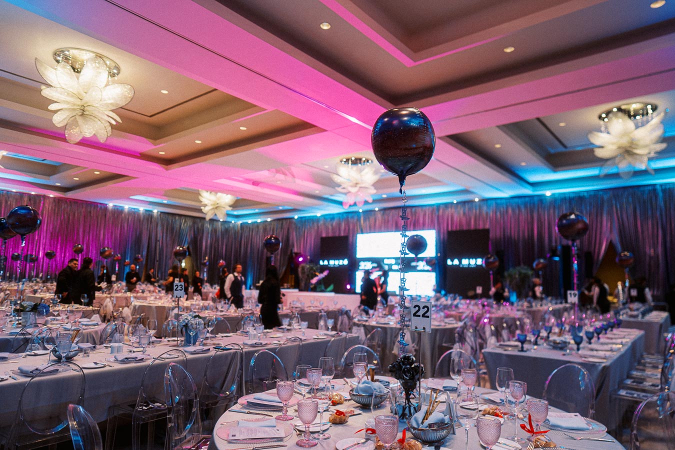 Elegant banquet hall decorated with pink and purple lighting, featuring round tables set with fine dining ware, glassware, and silverware, surrounded by transparent chairs. The ceiling is adorned with large flower-shaped chandeliers, and metallic balloons are suspended at each table, creating a festive atmosphere.