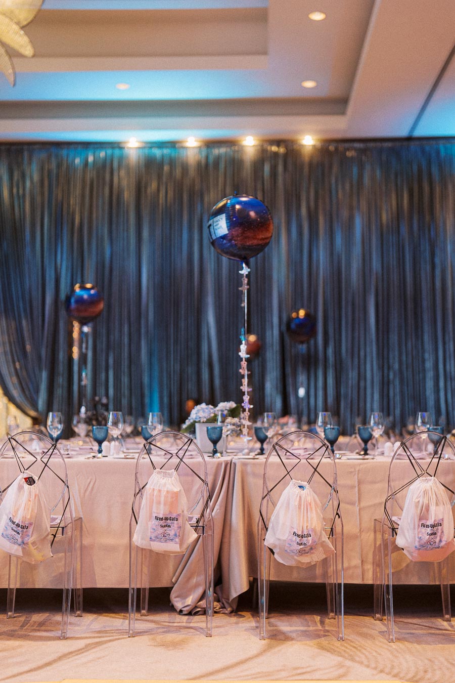 Elegant gala dinner setting with transparent chairs, draped with branded bags, around a long table adorned with blue tableware and decorative balloons in a modern venue.