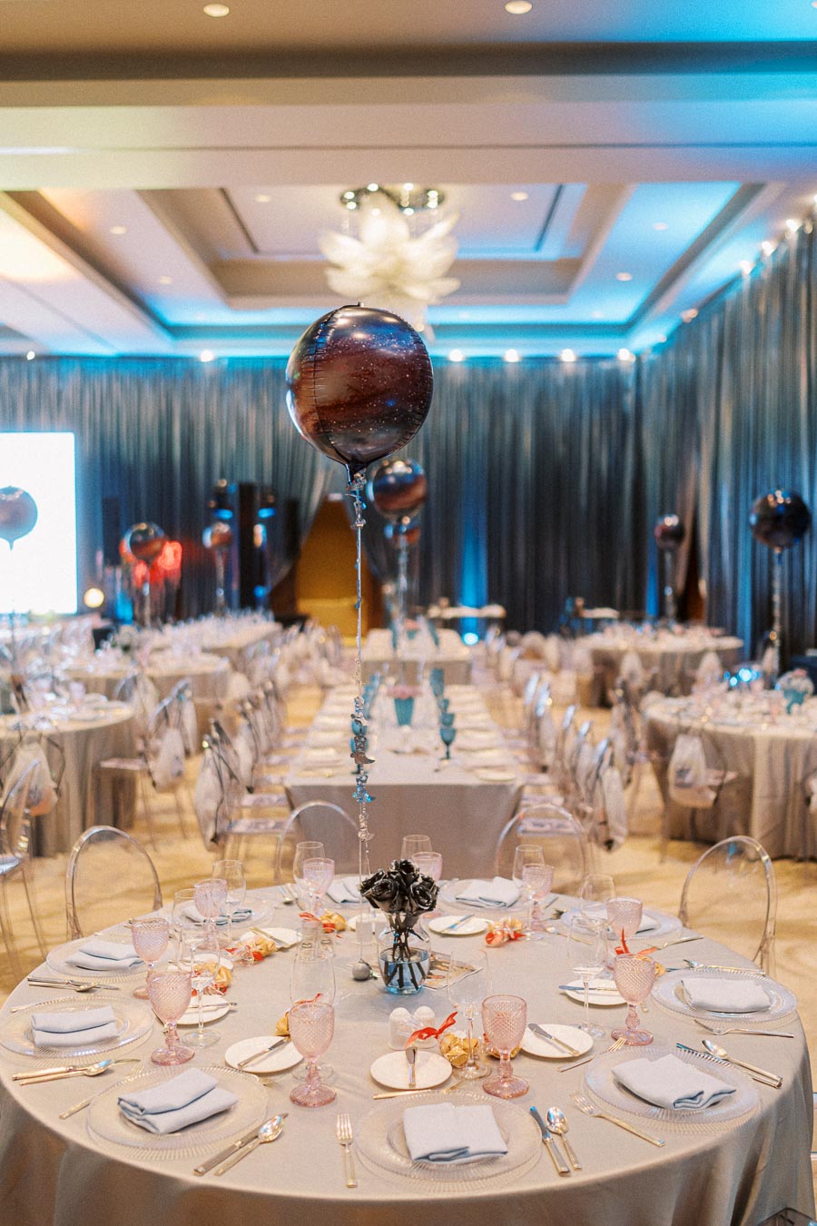 Elegant event setup with round tables draped in gray tablecloths, adorned with pink glassware and neatly folded napkins, in a spacious room with blue ambient lighting and decorative balloon centerpieces.