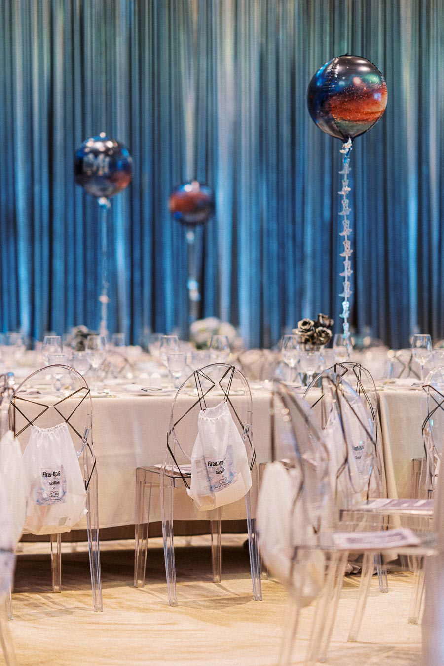 Elegant event dining setup with transparent chairs and white tablecloths, adorned with celestial-themed balloons and ambient blue lighting, conveying a sophisticated and modern atmosphere.