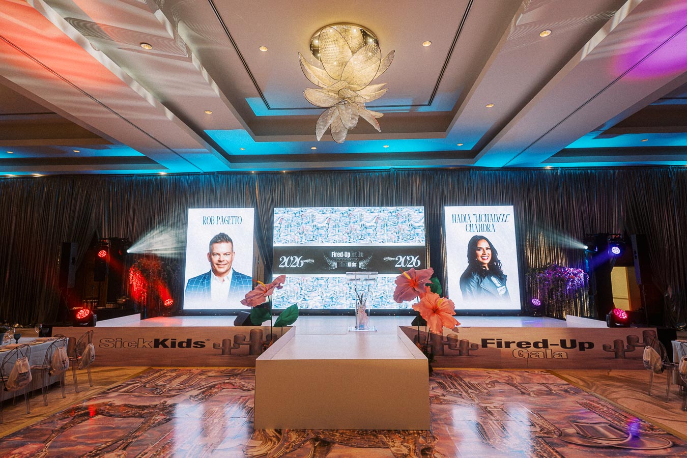 Elegant gala event setup with large screens displaying speakers at the SickKids Fired-Up Gala 2023. The stage is adorned with floral decor and dramatic lighting, creating a vibrant atmosphere for the occasion.