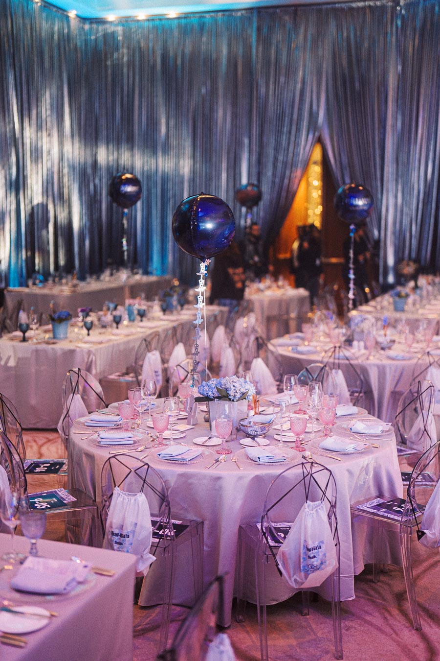 Elegant event venue with round tables draped in white tablecloths, accented by blue floral centerpieces and balloons. The dining setting features fine glassware and cutlery, surrounded by modern transparent chairs, creating a sophisticated atmosphere for a gala or special occasion.