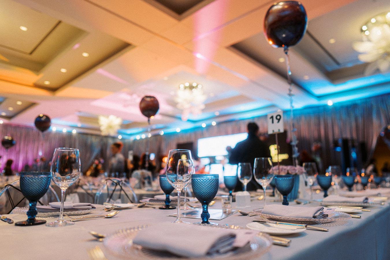 Elegant banquet hall setup with blue-tinted wine glasses, neatly arranged silverware, and balloons, creating a festive ambiance for a special event or celebration.