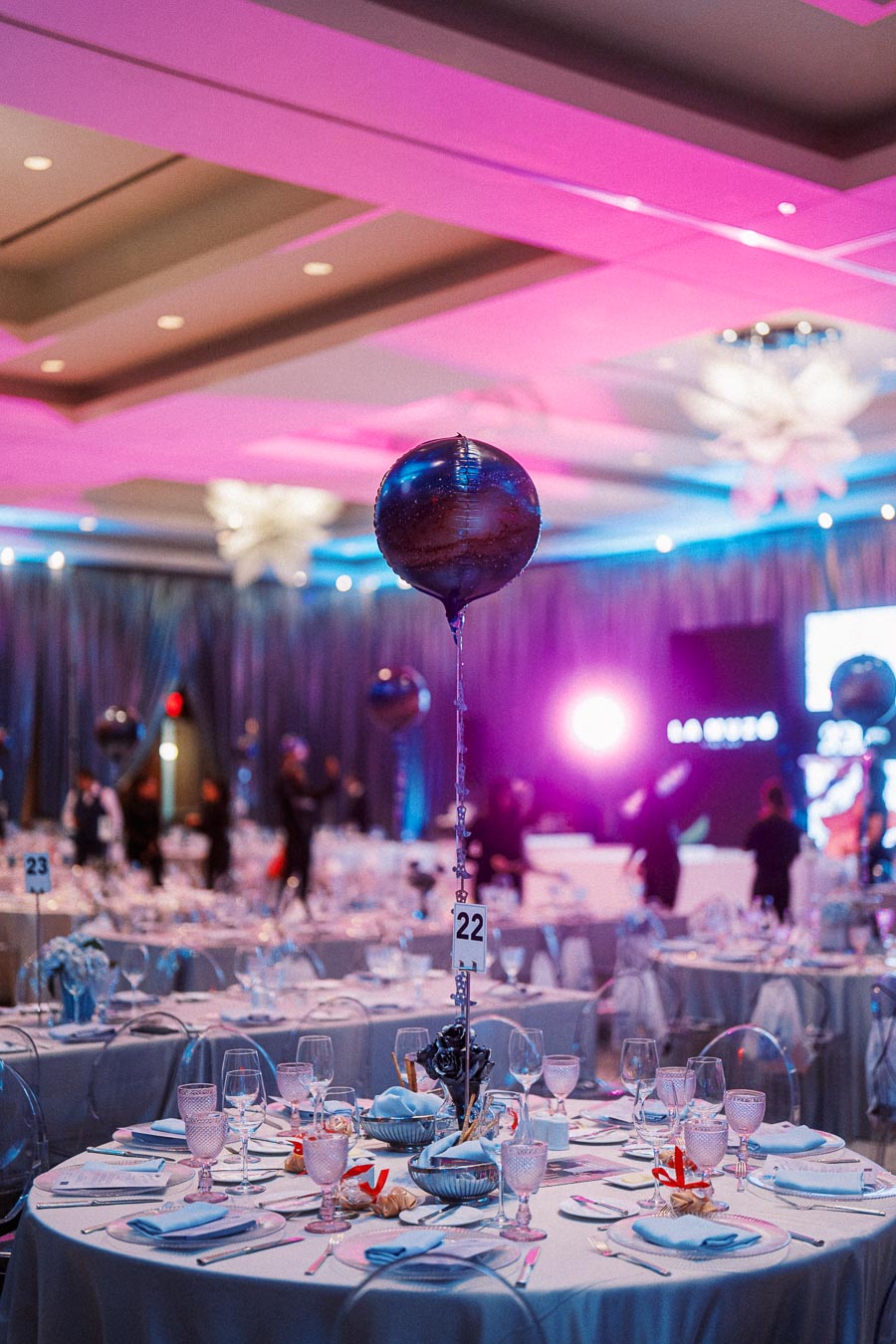 Elegant banquet hall setup with round tables adorned with white tablecloths, fine glassware, and decorative purple balloons, under ambient pink and blue lighting, creating a festive atmosphere.