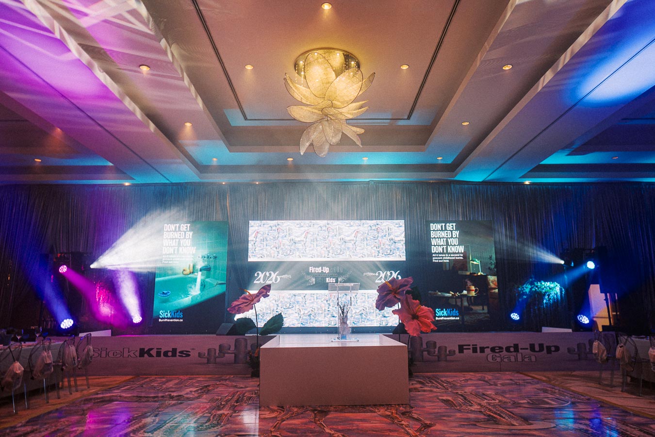 Luxurious event setup for the SickKids Fired-Up Gala 2026, featuring a vibrant stage with colorful lighting, large screen displays with messages, and elegant floral decorations in a lavish venue.