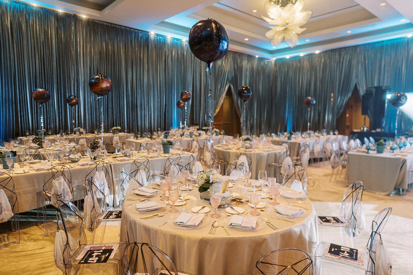 Elegant banquet hall setup for an event with round tables adorned with cream tablecloths, fine tableware, crystal glasses, and decorative centerpieces. Transparent chairs surround each table, enhanced by glossy balloon decor and soft ambient lighting.