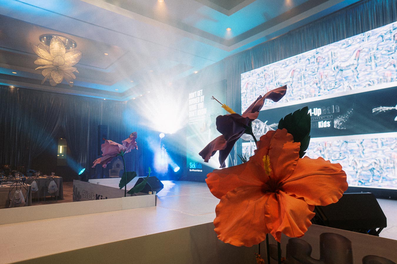 Elegant event stage with vibrant flower decorations, featuring a large digital screen displaying event information and dramatic lighting in a sophisticated venue setting.