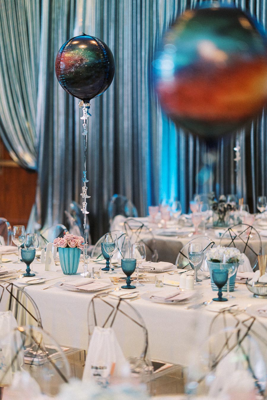 Elegant wedding reception setup featuring galaxy-themed balloons, blue glassware, and pastel floral centerpieces on a beautifully adorned table with a backdrop of shimmering silver curtains.