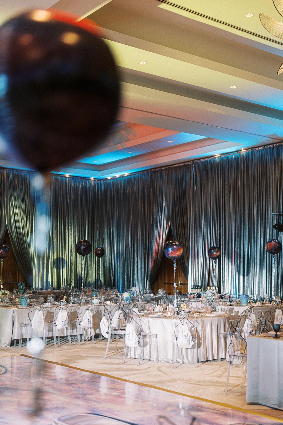 Elegant banquet hall with tables set for a formal event, featuring silver drapery, transparent chairs, and decorative balloons, creating a sophisticated ambiance.