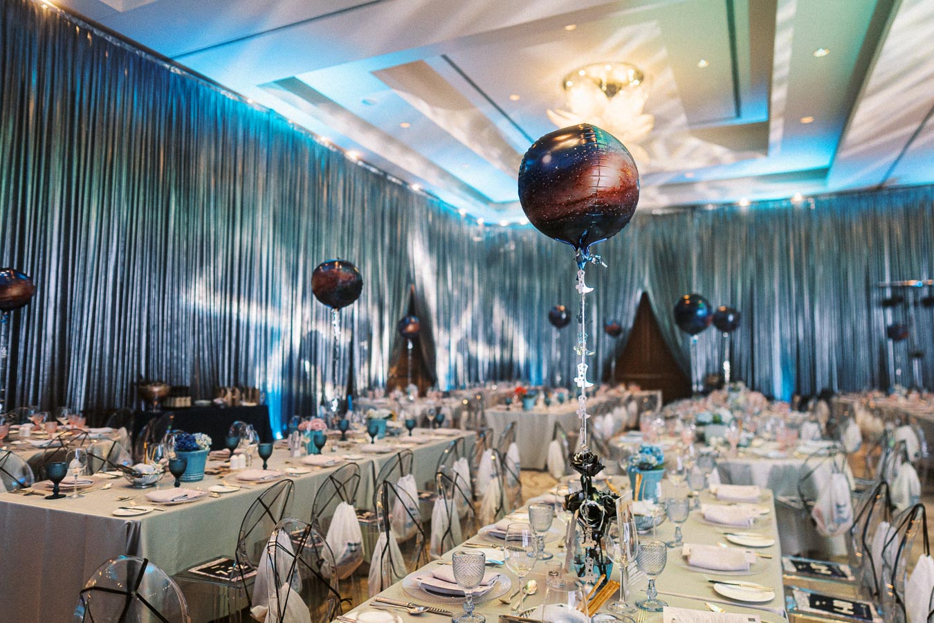 Elegant event venue with silver drapes and galaxy-themed decorations, including planetary balloons and beautifully set tables adorned with blue accents and glassware. Perfect setting for a sophisticated and modern celebration.