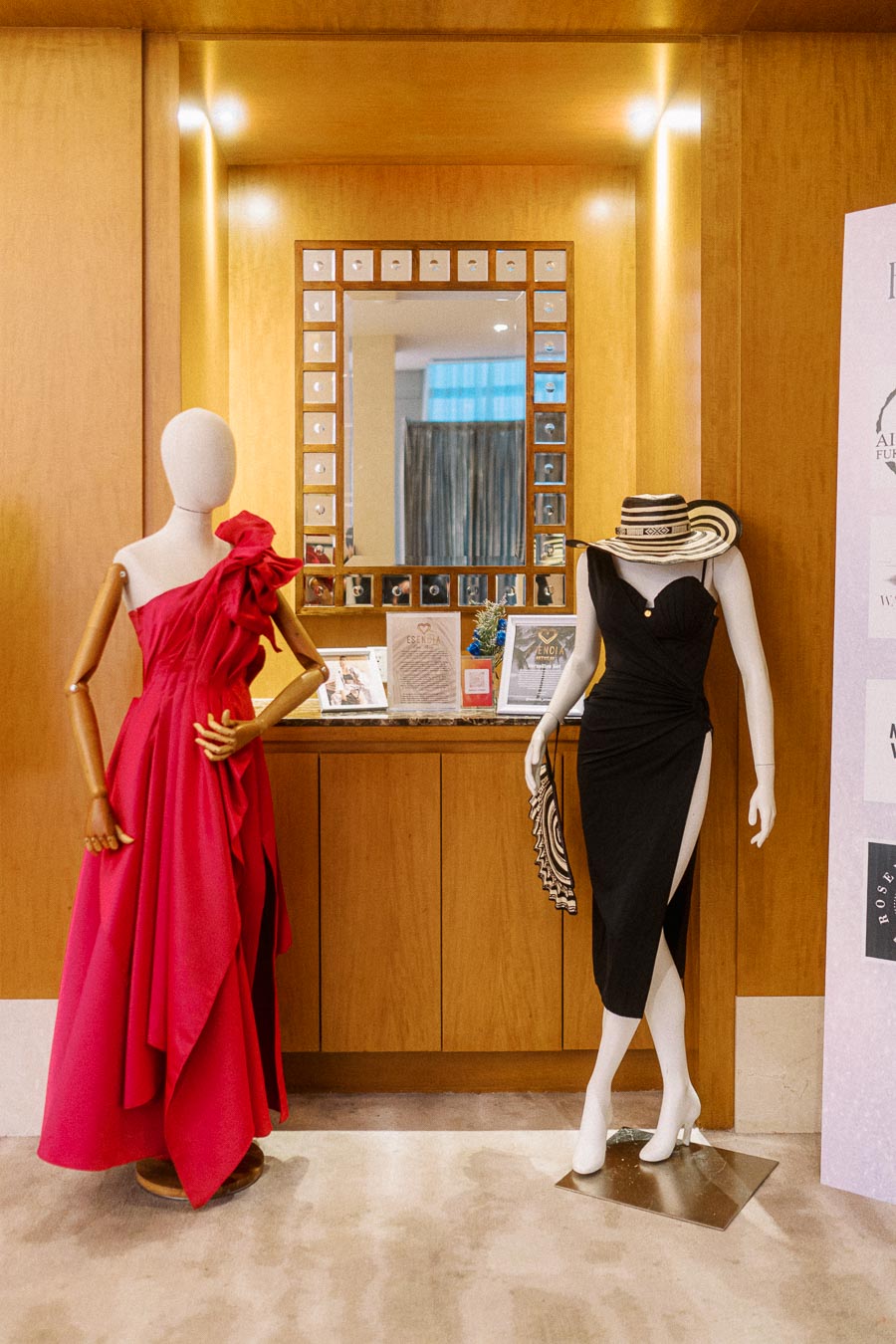 Fashion display featuring two mannequins, one in a vibrant red one-shoulder dress with ruffle details, and the other in an elegant black slit dress accessorized with a wide-brimmed hat, set against a wooden backdrop with a decorative mirror.