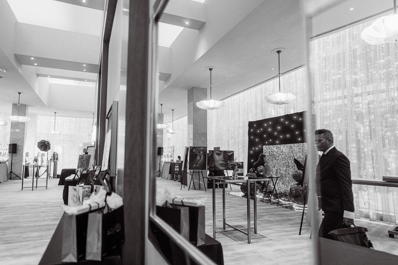 Black and white photograph of an elegant art exhibition space featuring paintings on easels, decorative lighting, and a sophisticated ambiance.