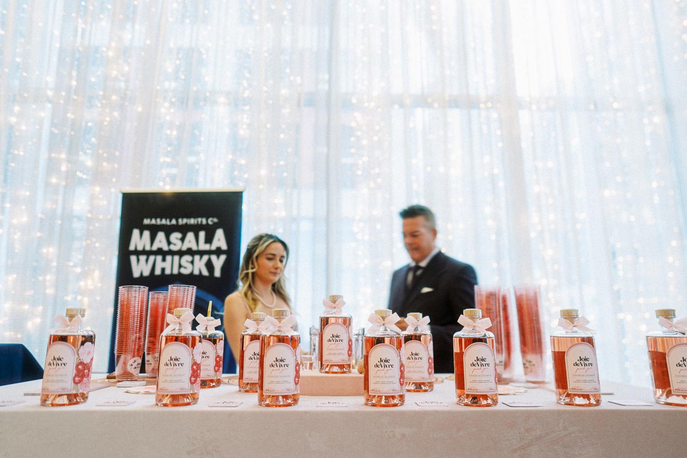 People standing behind a display table showcasing bottles of Joie de Vivre beverage with a Masala Whisky banner in the background, creating an elegant and festive atmosphere.