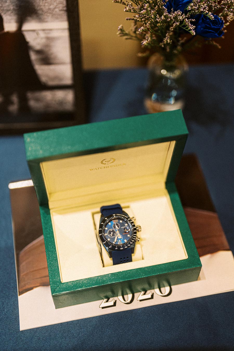 Luxury blue wristwatch in an open green Watchfinder box, placed on a blue surface with a calendar displaying the year 2020, and a vase with blue flowers in the background.