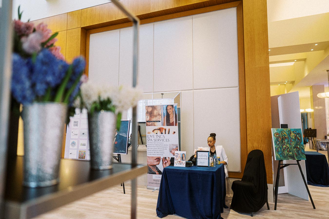 A woman sits at a table with a dark blue tablecloth featuring spa and beauty service promotional materials. There are artistic posters and banners around, along with colorful flower arrangements in metal buckets in the foreground, creating an inviting and professional atmosphere.