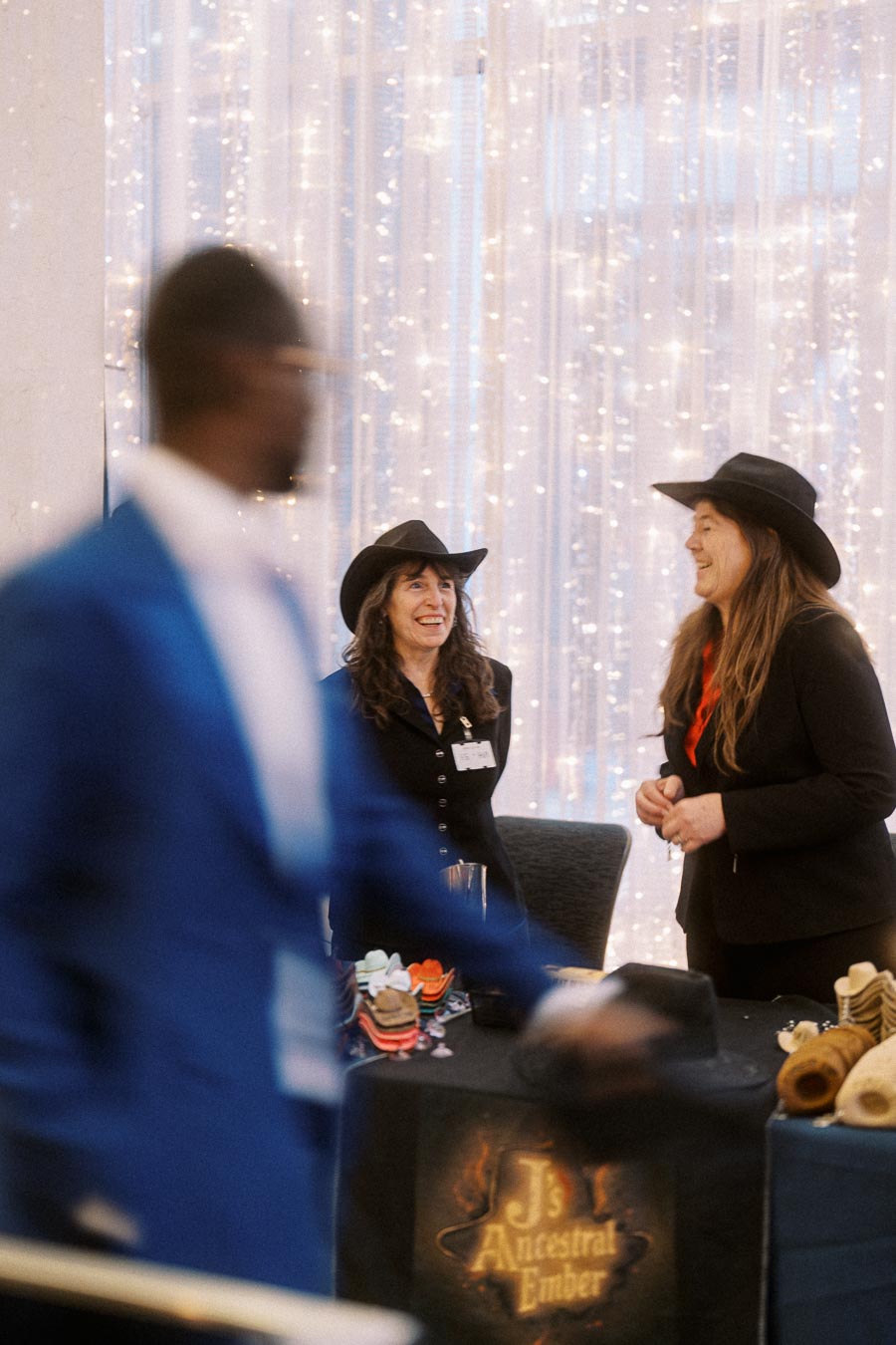 Two women wearing black cowboy hats smile while standing at a booth labeled J's Ancestral Ember at an indoor event, with a blur of a person in a blue suit in the foreground and festive lights in the background.