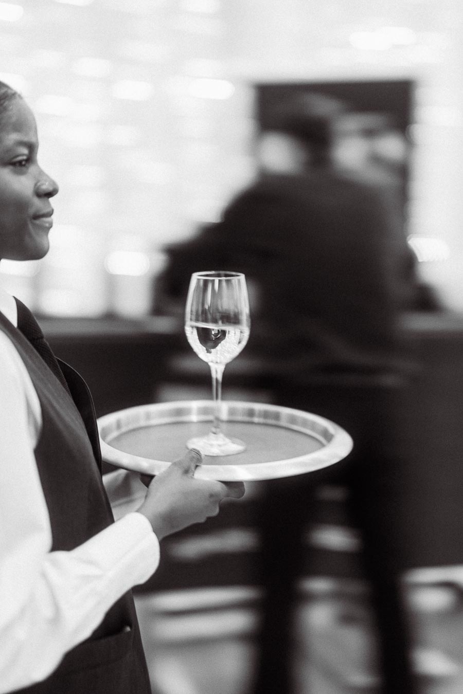 Black and white image of a professional waiter carrying a tray with a glass of water, set in an elegant restaurant environment.
