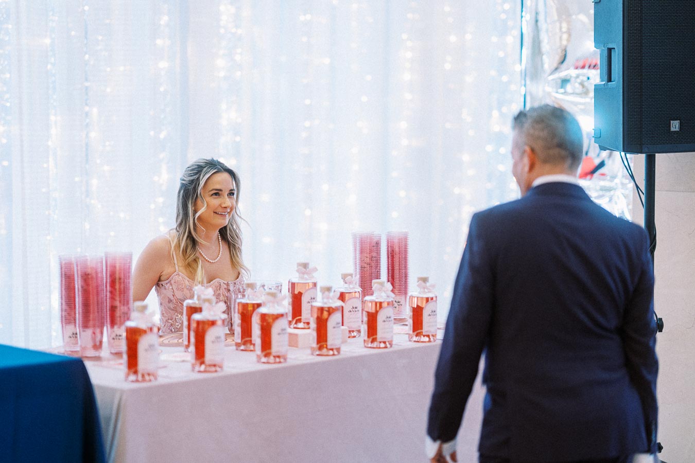 Young woman at a table with multiple bottles of pink liquid, smiling at a man in a suit at a festive event with twinkling lights in the background.