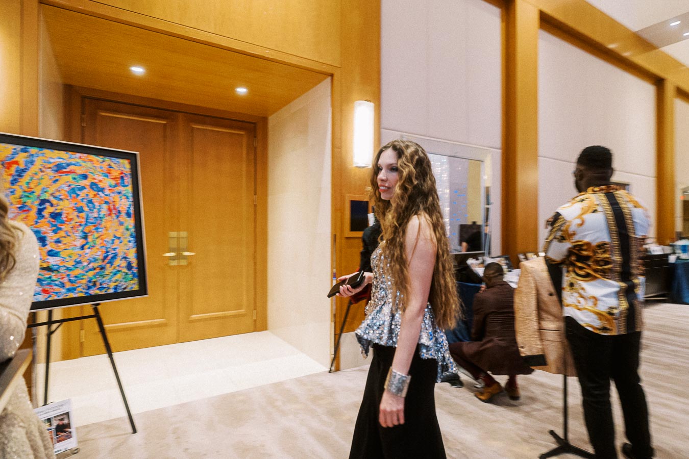 Elegant woman in a sparkling silver top walking through an art gallery with colorful abstract painting in the background, dressed attendees and upscale interior setting.