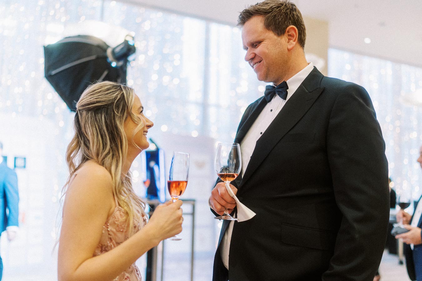 Elegant couple celebrating at a formal event with glasses of rosé champagne, man in a black tuxedo and woman in an evening gown, festive background with lights.