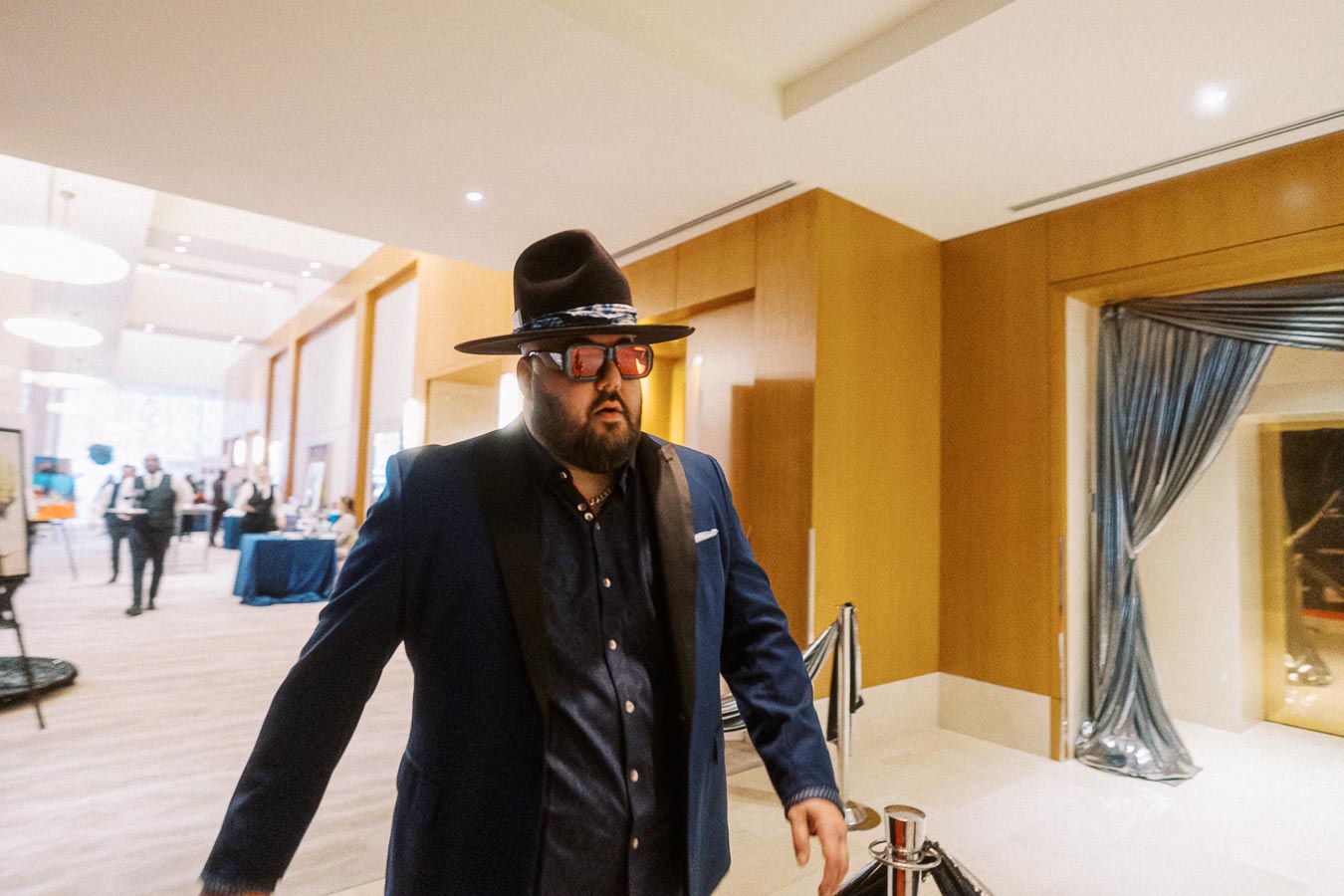 A stylish man in a hat and sunglasses walks through a modern, well-lit lobby with wood-paneled walls and guests visible in the background, conveying a sense of sophistication and event atmosphere.
