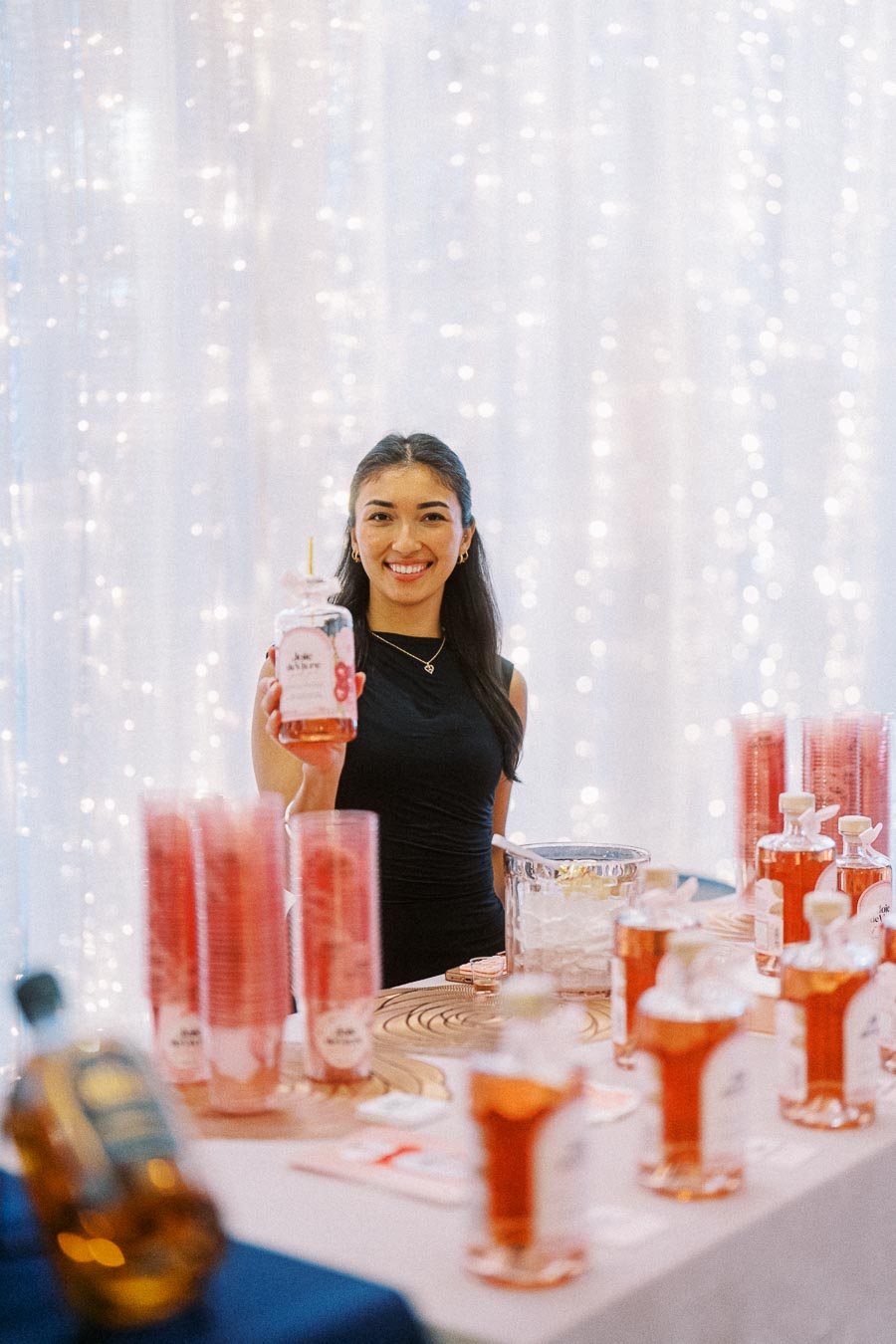 Smiling woman holding a bottle of rose syrup at a decorated beverage booth with fairy lights background.