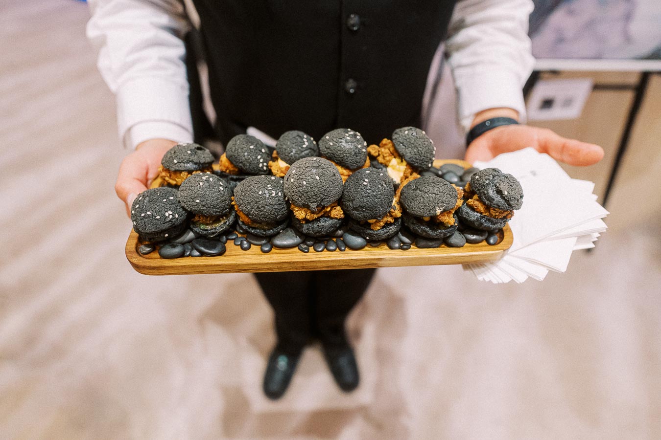 A person holding a wooden tray filled with gourmet black macarons topped with sesame seeds and filled with a savory mixture, showcasing an elegant catering presentation.