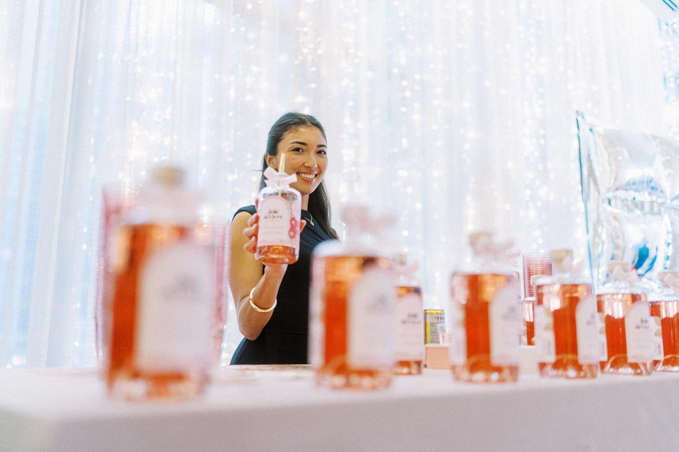 Smiling woman showcasing a rose-colored beverage bottle at a festive event with a backdrop of twinkling lights and a table filled with similar bottles.