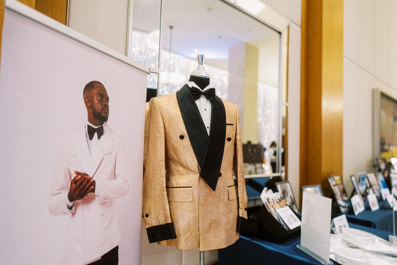 Exquisite gold textured tuxedo with black lapels displayed on a mannequin alongside a promotional banner featuring a man in a white suit with black bow tie, elegant fashion display in a modern showroom.
